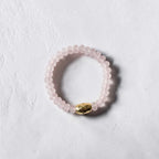 A beaded bracelet featuring light pink translucent round beads and a single polished gold oval accent bead, displayed on a textured white surface.