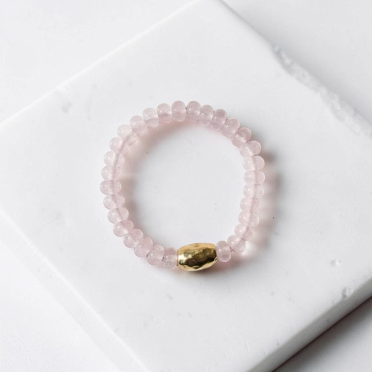 A pink beaded stretch bracelet with a hammered gold accent bead, displayed on a white marble block.