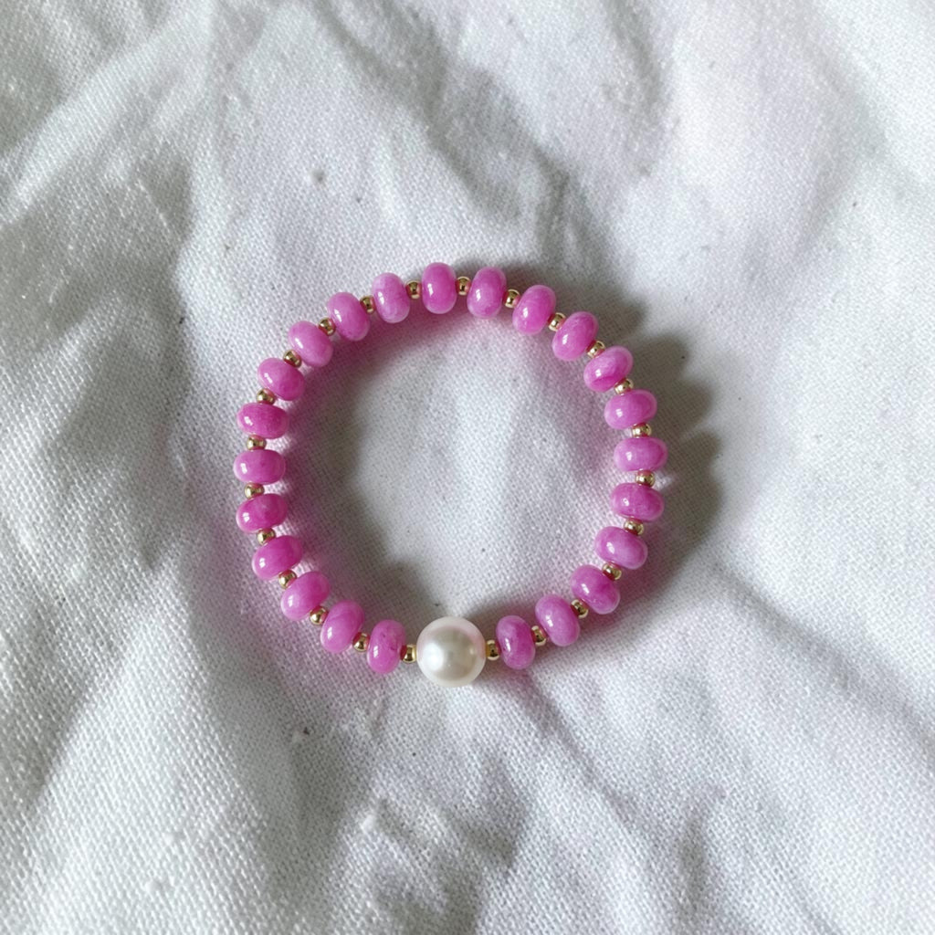 A pink beaded stretch bracelet with a single white pearl accent, displayed on a textured white fabric background.