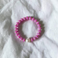 A pink beaded stretch bracelet with a single white pearl accent, displayed on a textured white fabric background.