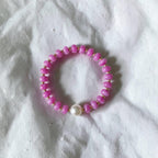 A pink beaded stretch bracelet with a single white pearl accent, displayed on a textured white fabric background.