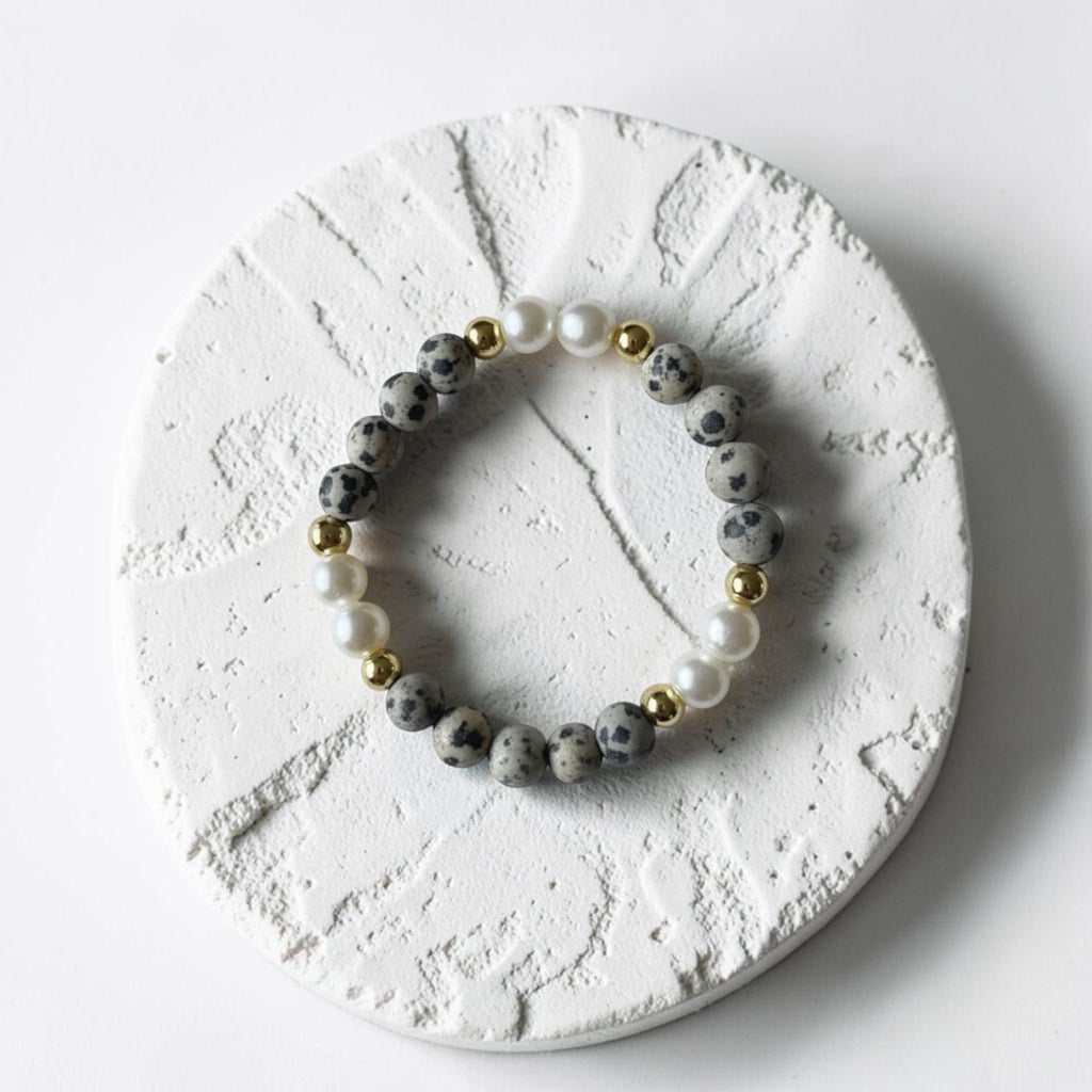 A beaded stretch bracelet featuring gray and black spotted beads, white pearl-like beads, and small gold spacer beads, displayed on a textured round white stone coaster.