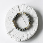 A beaded stretch bracelet featuring gray and black spotted beads, white pearl-like beads, and small gold spacer beads, displayed on a textured round white stone coaster.