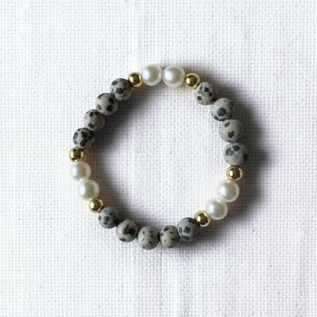 A beaded stretch bracelet featuring gray and black speckled stone beads, white faux pearls, and small gold spacer beads, displayed on a textured white fabric background.