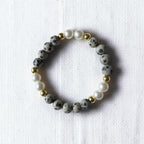 A beaded stretch bracelet featuring gray and black speckled stone beads, white faux pearls, and small gold spacer beads, displayed on a textured white fabric background.