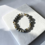 A beaded stretch bracelet with gray and white spotted beads, white faux pearls, and small gold spacer beads, displayed on a white stone tile under direct sunlight.