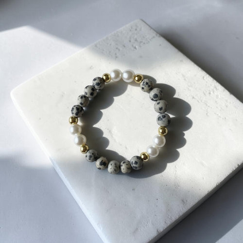 A beaded stretch bracelet with gray and white spotted beads, white faux pearls, and small gold spacer beads, displayed on a white stone tile under direct sunlight.