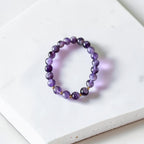 A beaded stretch bracelet made of polished purple gemstone beads with small gold spacer accents, displayed on a white textured stone tile.
