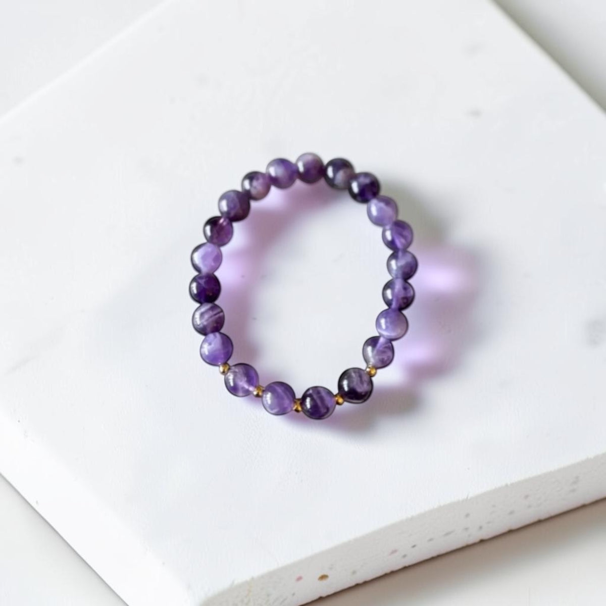 A beaded stretch bracelet made of polished purple gemstone beads with small gold spacer accents, displayed on a white textured stone tile.