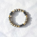 A beaded stretch bracelet featuring cream and black speckled stone beads, dark gray beads, and small polished gold spacer beads, displayed on a textured white surface.