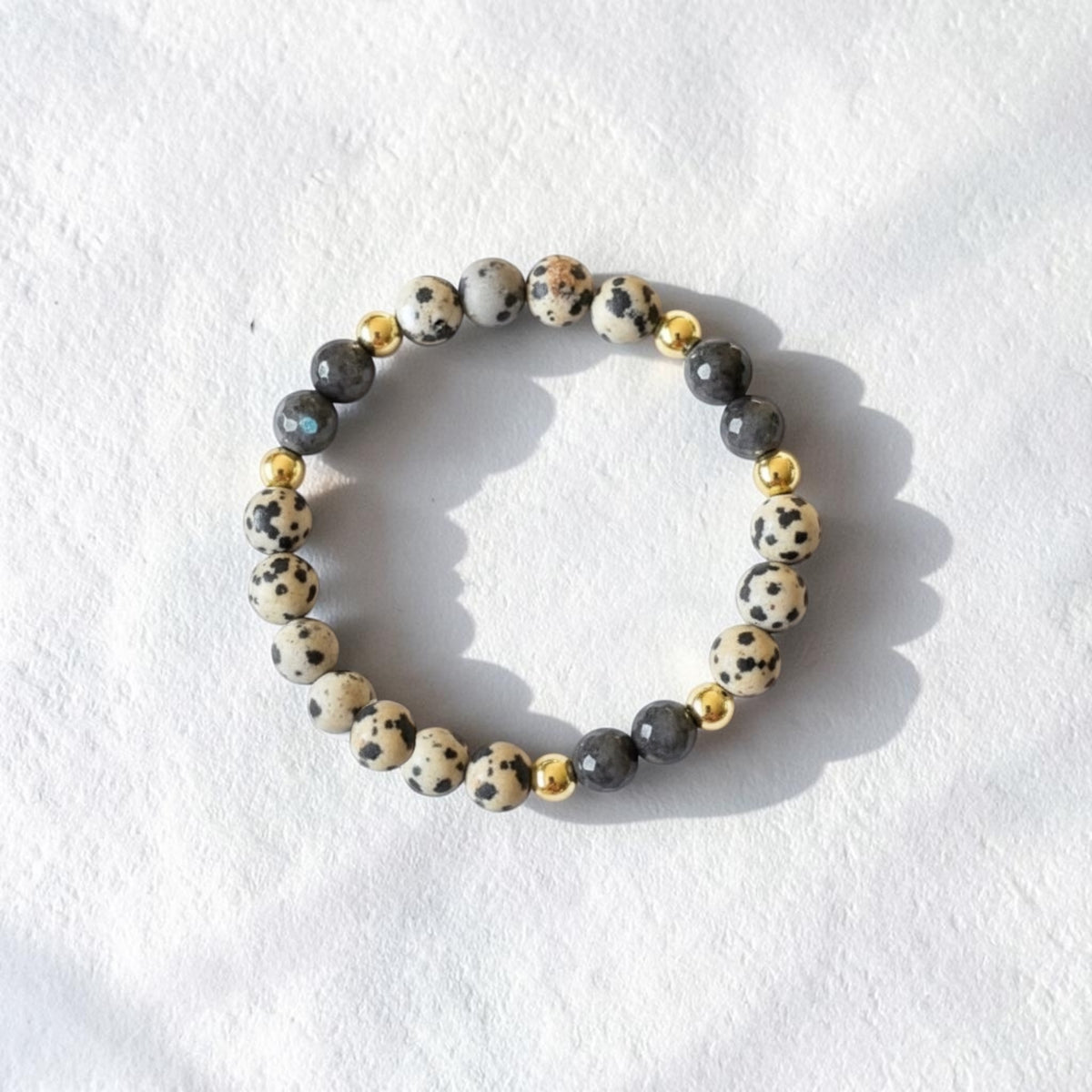 A beaded stretch bracelet featuring cream and black speckled stone beads, dark gray beads, and small polished gold spacer beads, displayed on a textured white surface.