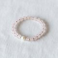 A delicate beaded stretch bracelet featuring light pink faceted beads, small gold spacer rings, and a single white pearl bead, displayed on a textured white fabric background.