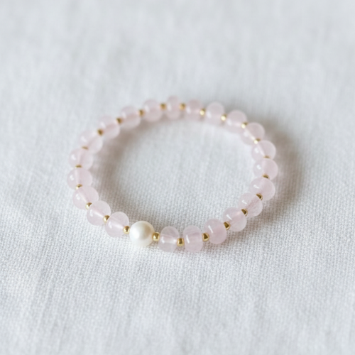 A delicate beaded stretch bracelet featuring light pink faceted beads, small gold spacer rings, and a single white pearl bead, displayed on a textured white fabric background.
