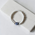 A white beaded stretch bracelet with a central blue and white ceramic fan-shaped bead, displayed on a white stone slab.
