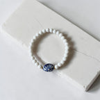 A white beaded stretch bracelet with a central blue and white ceramic fan-shaped bead, displayed on a white stone slab.