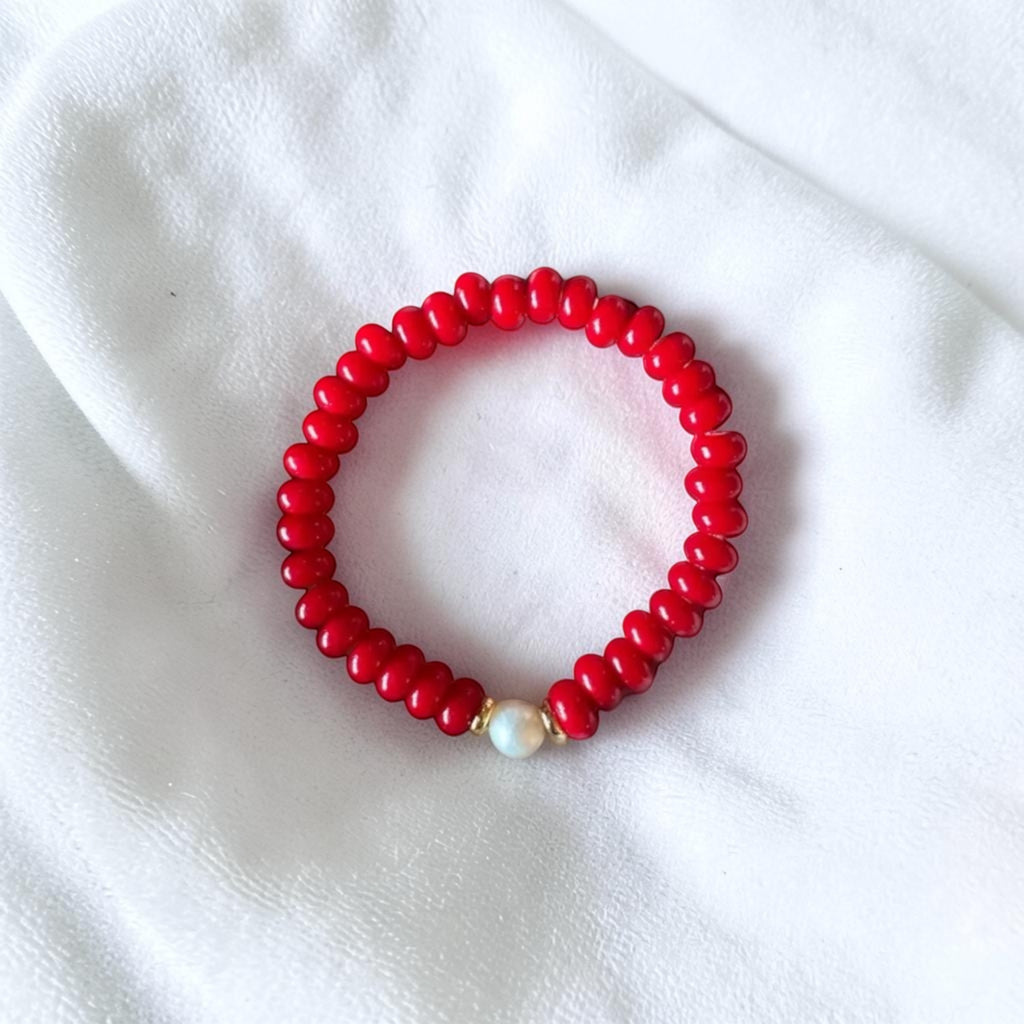A red beaded bracelet with a single white pearl accent and a small gold spacer, displayed on a soft white fabric background.