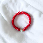 A red beaded bracelet with a single white pearl accent and a small gold spacer, displayed on a soft white fabric background.