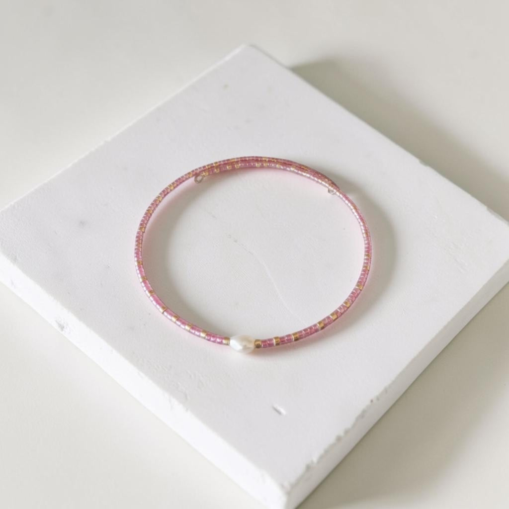 A delicate pink beaded bangle bracelet with a small white pearl accent, displayed on a white textured square block.