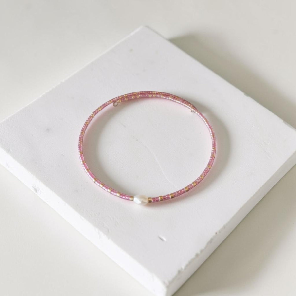 A delicate pink beaded bangle bracelet with a small white pearl accent, displayed on a white textured square block.