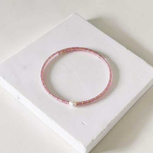 A delicate pink beaded bangle bracelet with a small white pearl accent, displayed on a white textured square block.