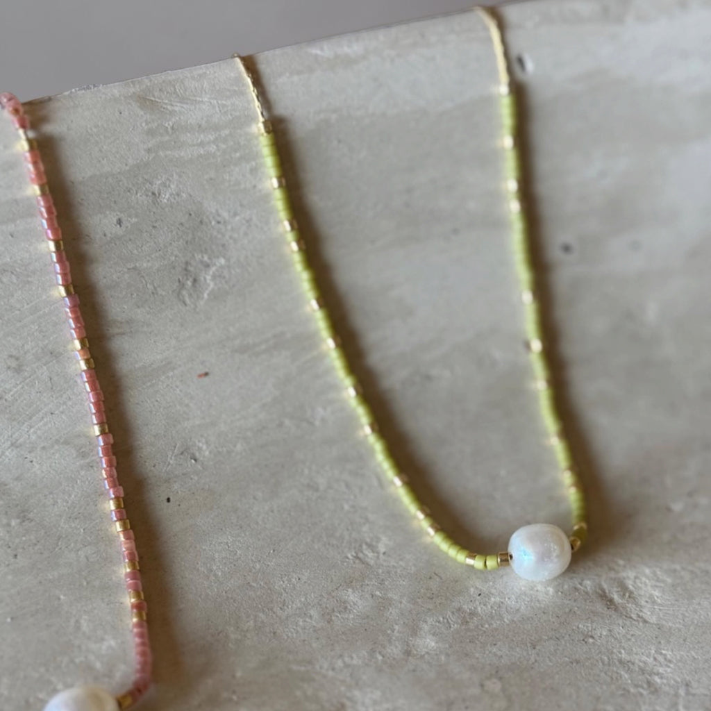 Two delicate beaded necklaces displayed on a textured light gray surface. One necklace is pink with a white pearl bead, and the other is pale yellow-green with a white pearl bead.