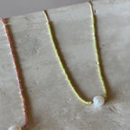 Two delicate beaded necklaces displayed on a textured light gray surface. One necklace is pink with a white pearl bead, and the other is pale yellow-green with a white pearl bead.