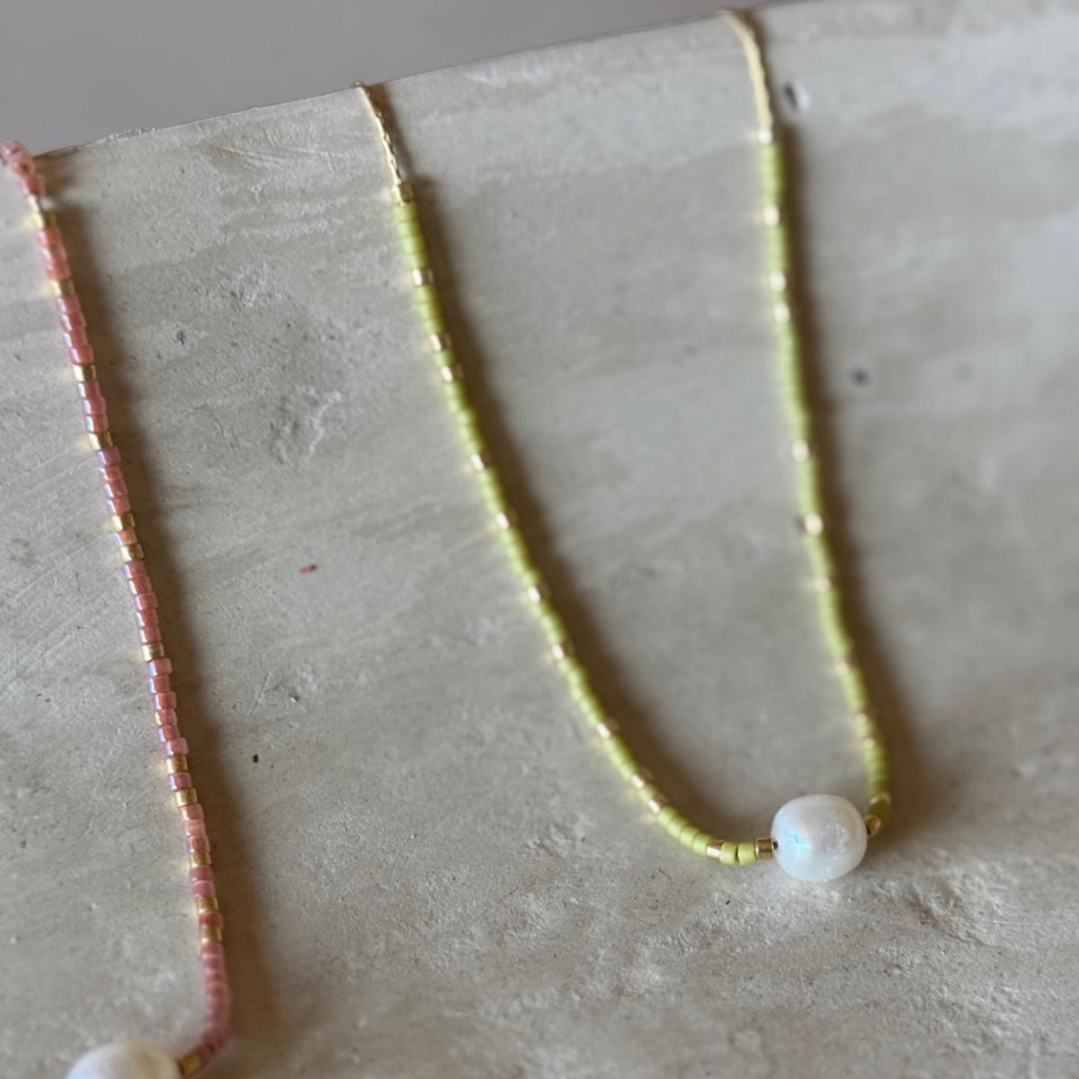 Two delicate beaded necklaces displayed on a textured light gray surface. One necklace is pink with a white pearl bead, and the other is pale yellow-green with a white pearl bead.