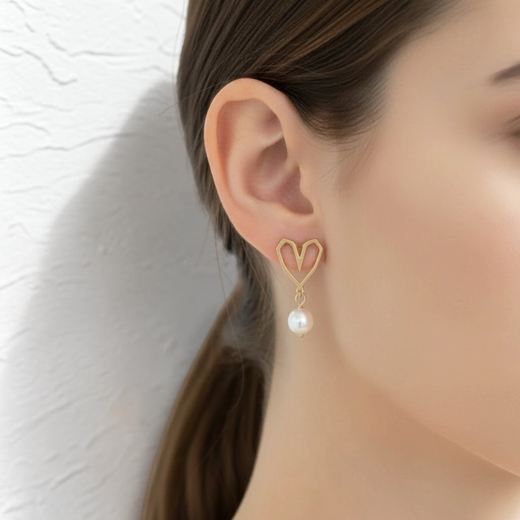 A close-up of a woman's ear wearing a gold heart-shaped dangle earring with a white pearl drop, set against a textured white wall.