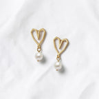 A pair of gold heart-shaped dangle earrings with white pearl drops, displayed on a soft, textured white fabric background.