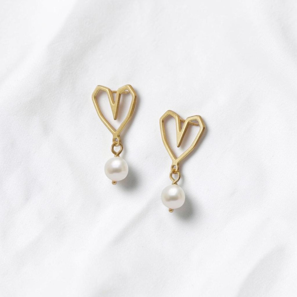 A pair of gold heart-shaped dangle earrings with white pearl drops, displayed on a soft, textured white fabric background.