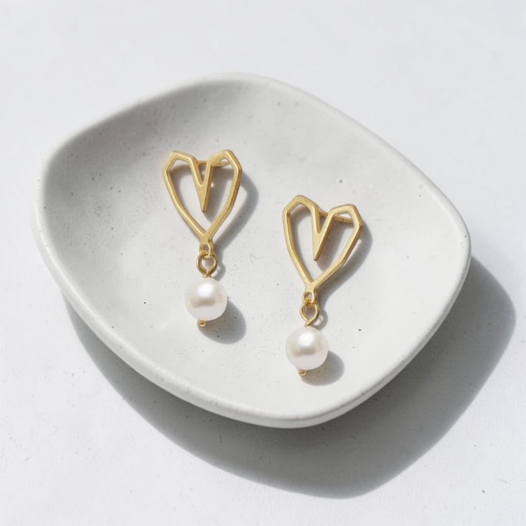 A pair of gold heart-shaped dangle earrings with white pearl drops, displayed on a small white ceramic dish against a light gray background.