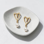 A pair of gold heart-shaped dangle earrings with white pearl drops, displayed on a small white ceramic dish against a light gray background.