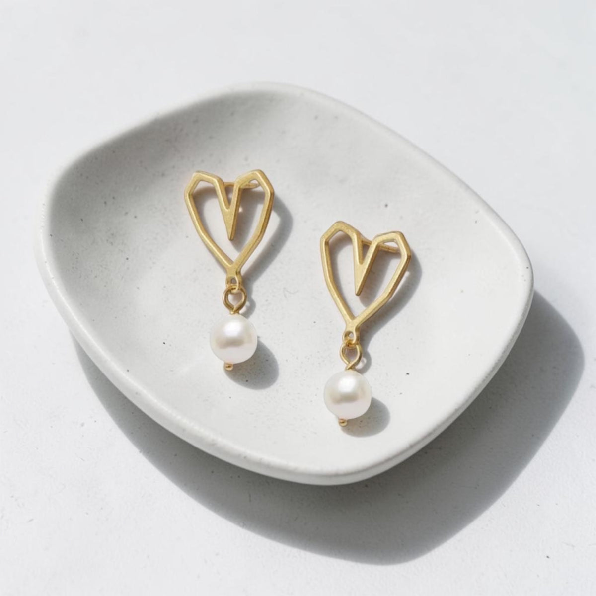 A pair of gold heart-shaped dangle earrings with white pearl drops, displayed on a small white ceramic dish against a light gray background.