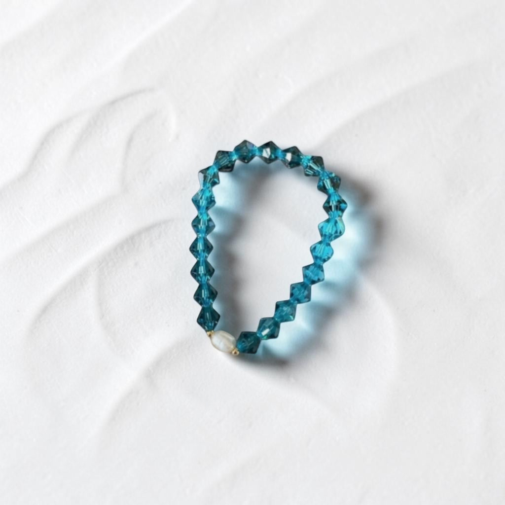 A beaded stretch bracelet made of faceted teal-blue beads, with a small white pearl accent and a tiny gold spacer, displayed on a textured white surface.
