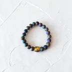 A beaded stretch bracelet featuring iridescent dark blue, green, and purple round beads, accented by a single textured gold-toned bead, displayed on a textured white surface.