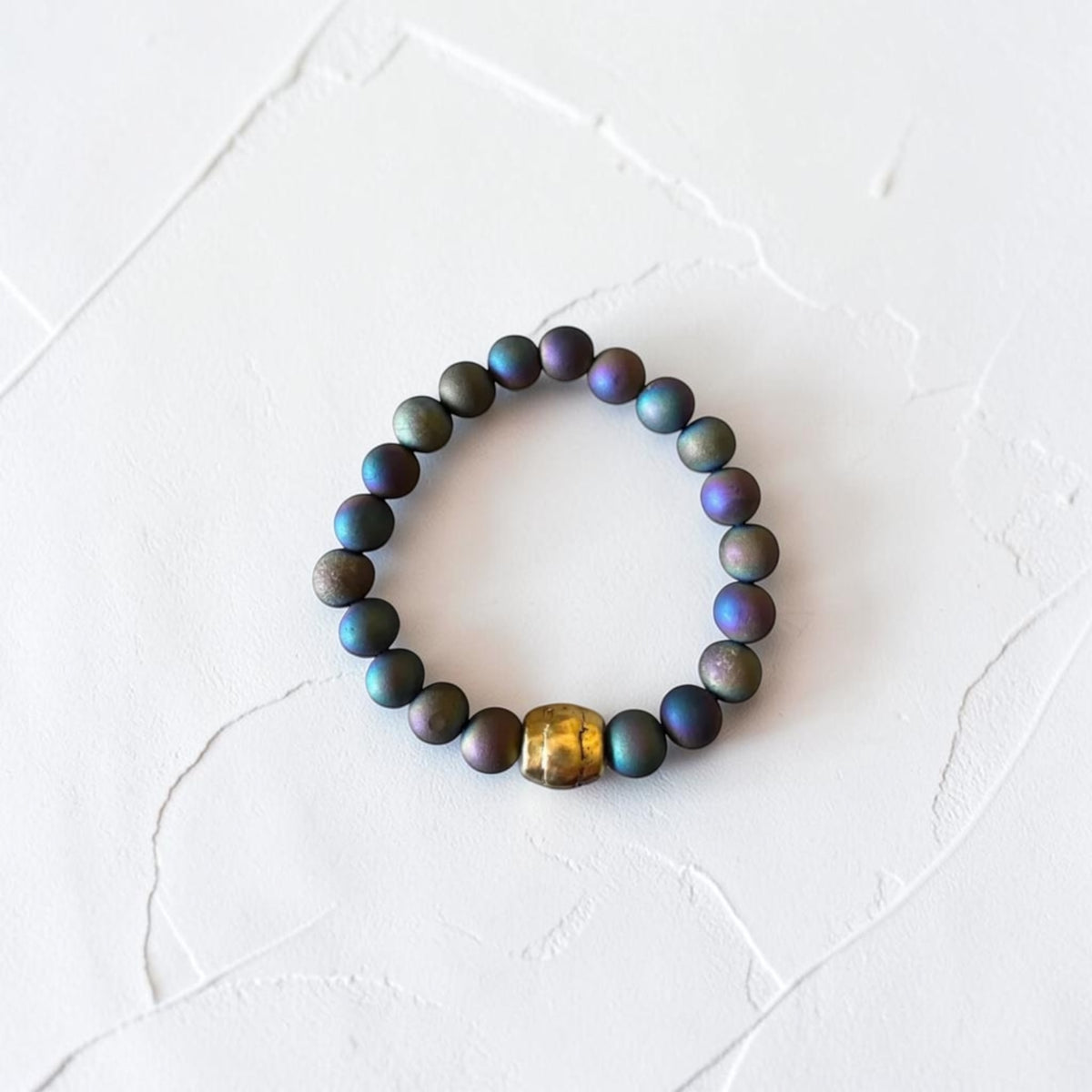 A beaded stretch bracelet featuring iridescent dark blue, green, and purple round beads, accented by a single textured gold-toned bead, displayed on a textured white surface.