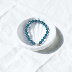 A beaded stretch bracelet with faceted teal-blue beads and a small white pearl accent, displayed on a white scalloped dish over a white fabric background.