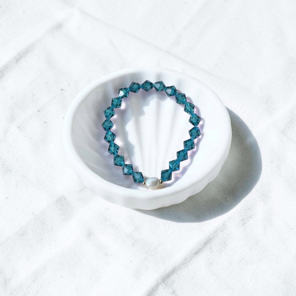 A beaded stretch bracelet with faceted teal-blue beads and a small white pearl accent, displayed on a white scalloped dish over a white fabric background.