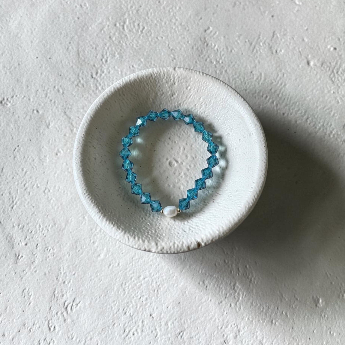 A beaded stretch bracelet with light blue faceted beads and a white pearl accent, displayed on a small textured white ceramic dish against a light gray background.