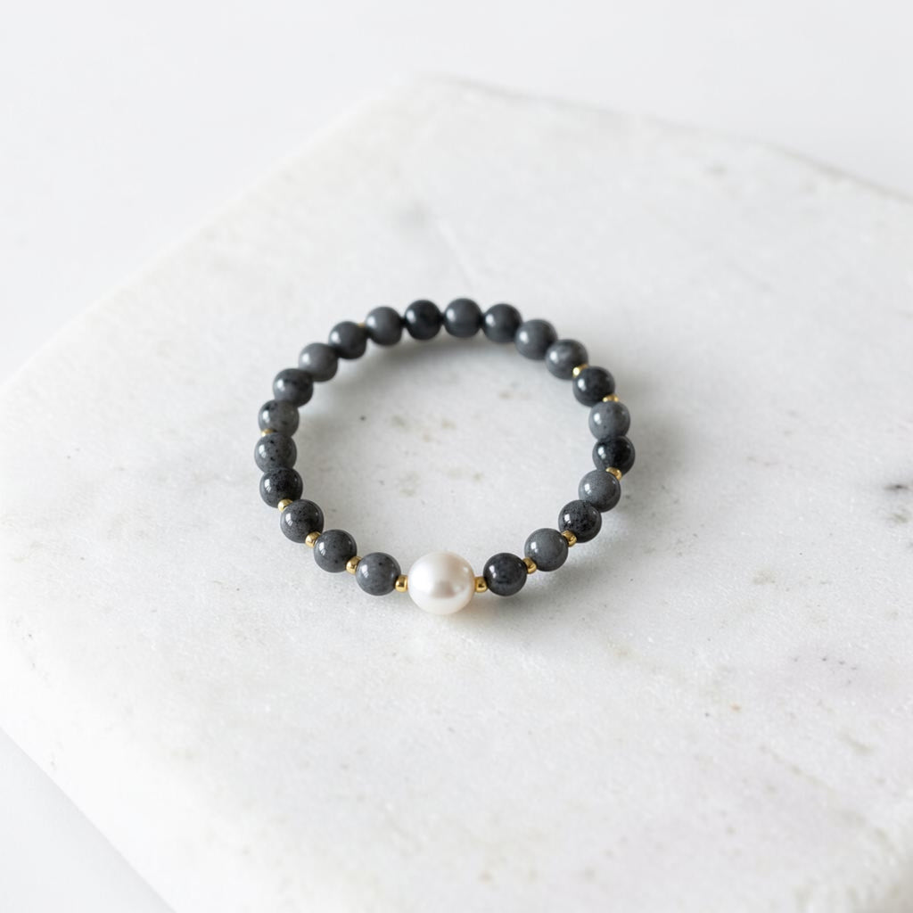 A beaded stretch bracelet featuring dark gray stone beads, small gold spacer beads, and a single white pearl accent, displayed on a white marble slab.