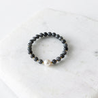 A beaded stretch bracelet featuring dark gray stone beads, small gold spacer beads, and a single white pearl accent, displayed on a white marble slab.