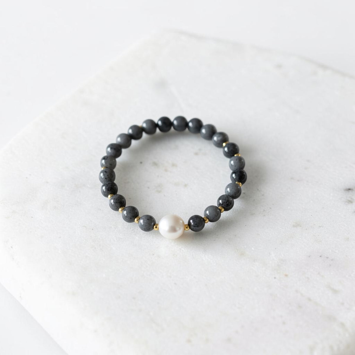 A beaded stretch bracelet featuring dark gray stone beads, small gold spacer beads, and a single white pearl accent, displayed on a white marble slab.