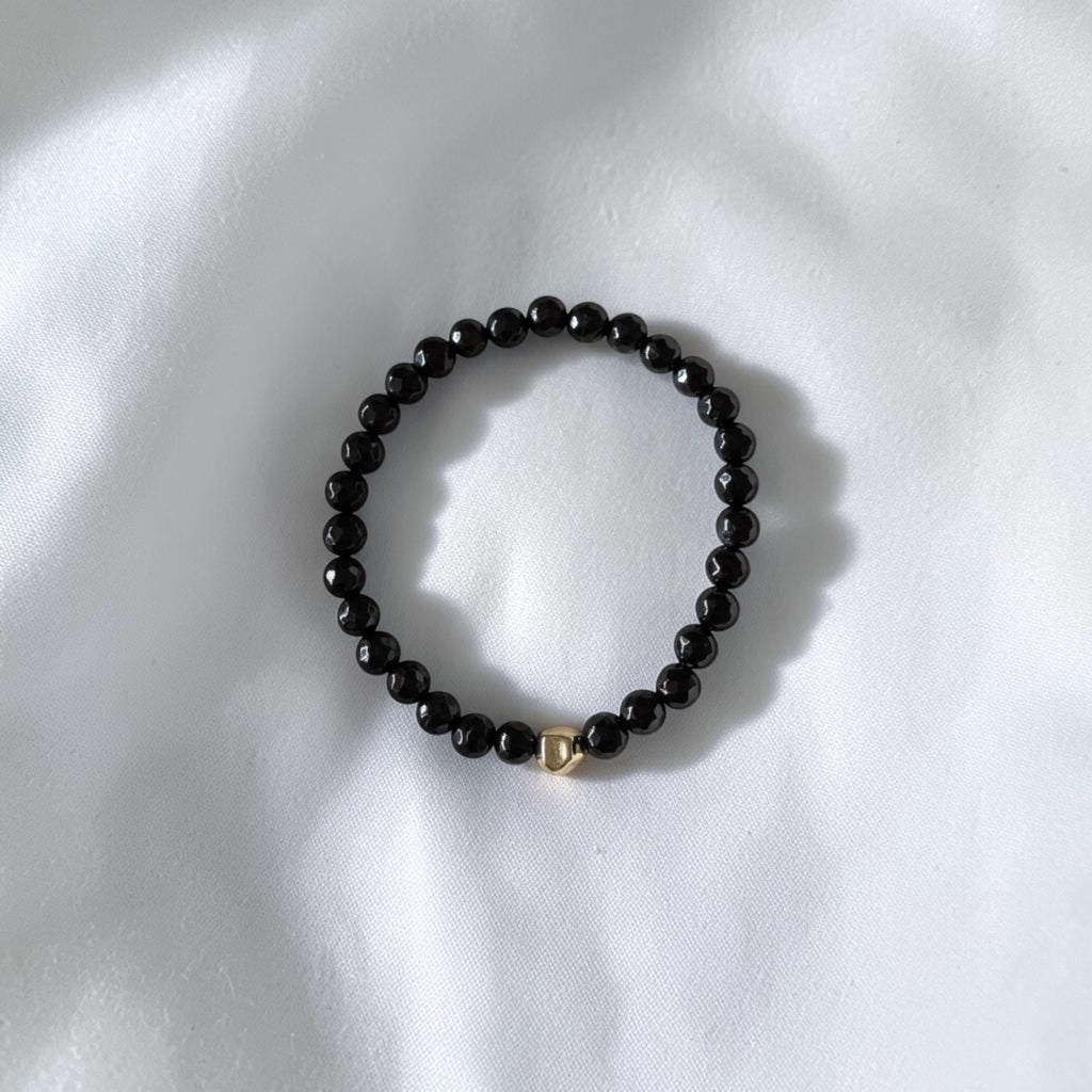 A beaded stretch bracelet made of small glossy black beads with a small gold-toned heart charm, resting on a white fabric background.