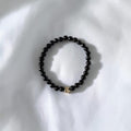 A beaded stretch bracelet made of small glossy black beads with a small gold-toned heart charm, resting on a white fabric background.