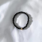 A beaded stretch bracelet made of small glossy black beads with a small gold-toned heart charm, resting on a white fabric background.