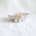 A pair of gold heart-shaped dangle earrings with white pearl drops, displayed on a small white ceramic dish over a textured white fabric background.