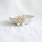 A pair of gold heart-shaped dangle earrings with white pearl drops, displayed on a small white ceramic dish over a textured white fabric background.