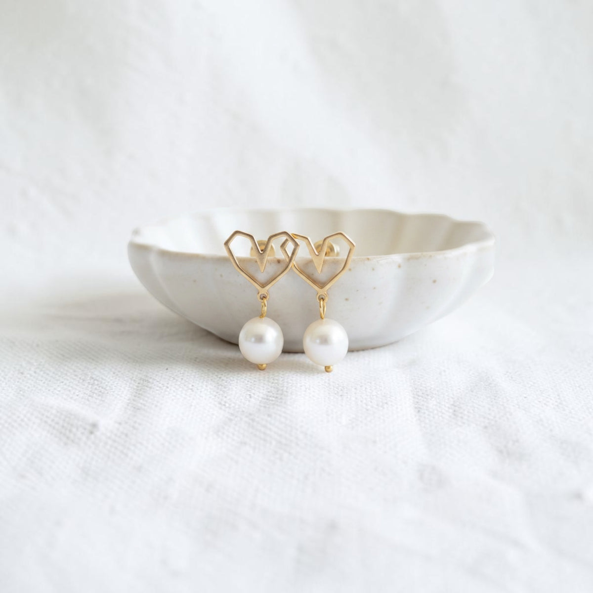 A pair of gold heart-shaped dangle earrings with white pearl drops, displayed on a small white ceramic dish over a textured white fabric background.