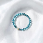 A beaded stretch bracelet featuring faceted light blue beads and a single white pearl accent, displayed on a soft, white textured fabric background.