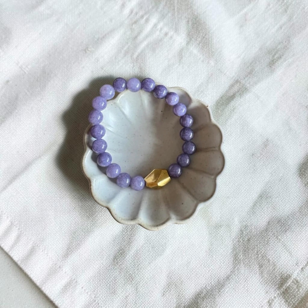 A beaded stretch bracelet with light purple round beads and a single gold accent bead, displayed on a small scalloped ceramic dish over a white fabric background.